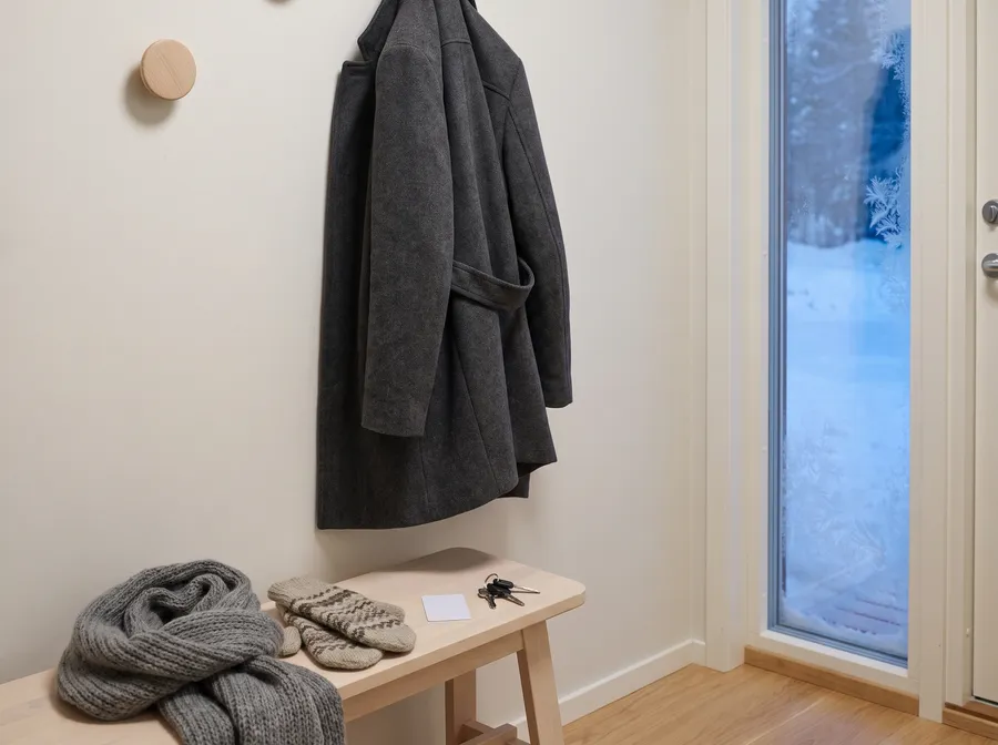 Generated scene: a charcoal wool coat on a wall peg, a grey scarf, mittens, a blank card and keys on a wooden bench, a frosted glass door to the right.